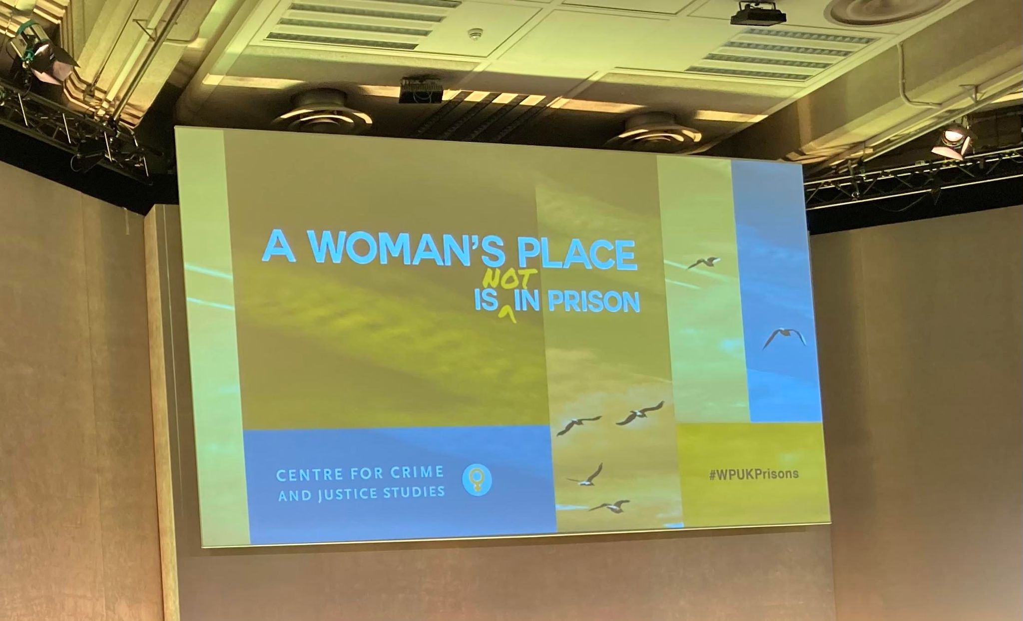 Photo shows screen above stage at #WPUKPrisons meeting - A Woman's Place is not in prison - shades of yellow, grey